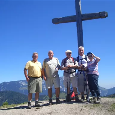 Eine Gruppe von fünf Wanderern steht vor einem großen Holzkreuz in den Bergen. Die Umgebung zeigt eine beeindruckende Landschaft mit Bergen und klarem Himmel.