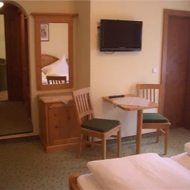 Ein gemütliches Hotelzimmer mit zwei Betten, einem Tisch, Stühlen und einem Fernseher. Im Hintergrund ist ein Torbogen zu sehen, das in einen anderen Raum führt.