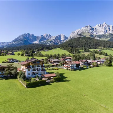 Eine malerische Landschaft mit grünen Wiesen und charmanten Gebäuden. Im Hintergrund erheben sich beeindruckende Berge unter klarem Himmel.