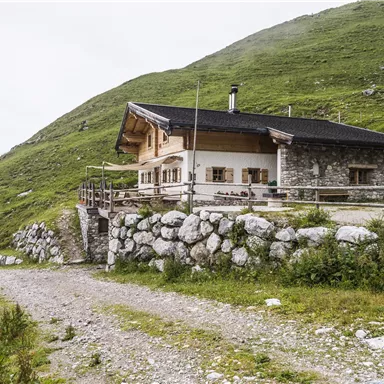 Eine gemütliche Hütte in den Bergen, umgeben von üppigem Grün. Der Weg zur Hütte ist von Steinen und Pflanzen gesäumt.