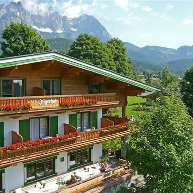 Ein charmantes Holzhaus mit Blumenkästen und vielen Fenstern. Im Hintergrund erheben sich majestätische Berge unter einem klaren Himmel.
