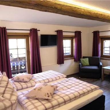 A cozy bedroom with two beds and checked bed covers. The windows are decorated with red curtains, and a small television is mounted on the wall.