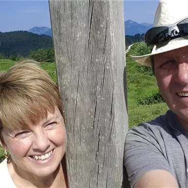 A cheerful couple poses in front of a wooden post in a beautiful, green landscape. In the background, gentle hills and mountains can be seen.