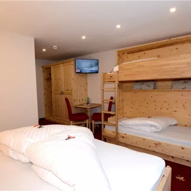 Ein gemütliches Zimmer mit einem Doppelbett und einem Etagenbett aus Holz. Im Hintergrund sind ein Fernseher und ein Tisch mit Stühlen zu sehen.