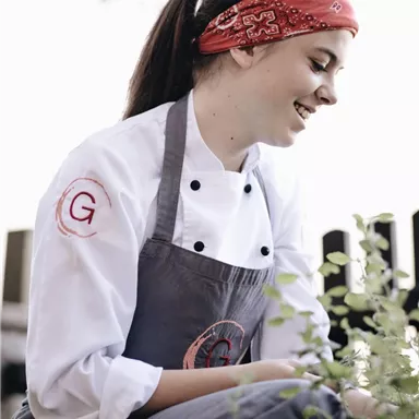 Eine Köchin mit einem roten Bandana kniet und pflückt Kräuter. Sie trägt eine weiße Kochjacke und ist von Pflanzen umgeben.