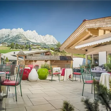 Eine gemütliche Terrasse mit Tischen und Stühlen, umgeben von einer landschaftlichen Kulisse mit Bergen im Hintergrund. Der Himmel ist klar, und die Atmosphäre wirkt einladend und entspannend.