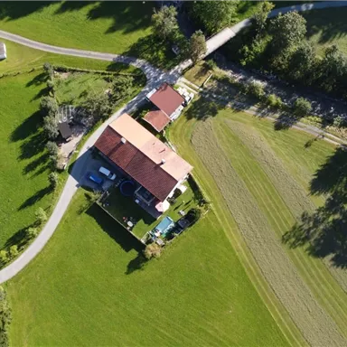 An aerial view of a rural area with several houses and green meadows. The paths are clearly visible and surrounded by trees.