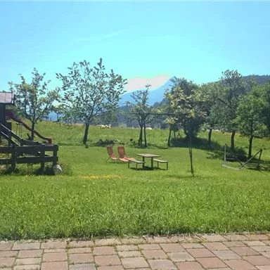 A green meadow with trees and a bench is in the foreground. In the background, mountains and a blue sky can be seen.