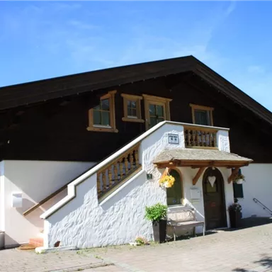 Ein charmantes Haus im traditionellen Stil mit einem Holzobergeschoss. Der Eingangsbereich ist freundlich gestaltet, umgeben von grüner Vegetation.