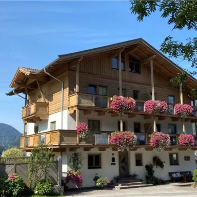 Ein charmantes Holzhaus im alpinen Stil mit Balkonen, die mit bunten Blumen geschmückt sind. Im Hintergrund sind sanfte Berge und ein klarer blauer Himmel zu sehen.