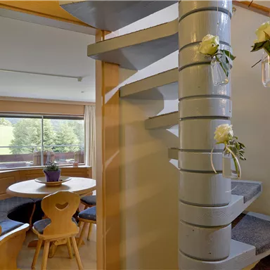 A bright entrance area with a curved staircase and flowers in glass containers. In the background, a cozy dining area with a window and a view of the outdoors can be seen.
