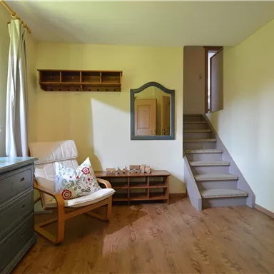 Ein helles Zimmer mit einer gemütlichen Sitzgelegenheit und einem Holzregal. Eine Treppe führt in den oberen Bereich, während ein großes Fenster natürliches Licht hereinlässt.