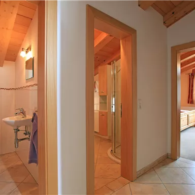 A bright hallway with a wooden ceiling and two doors. On the left side, a bathroom with a shower and sink is visible.