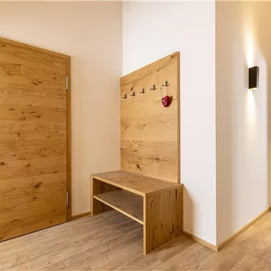 Ein heller Flur mit einem Holztür und einem Holzbank. An der Wand hängen Garderobenhaken und ein kleines Herzdekoration.