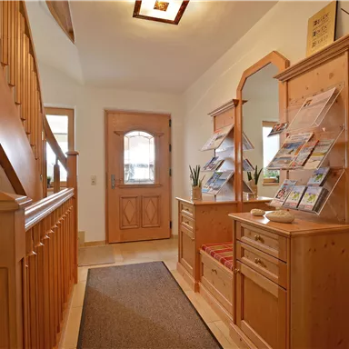 An inviting hallway with wooden interior and a staircase. On the left side, there is a piece of furniture with informational brochures and a plant.