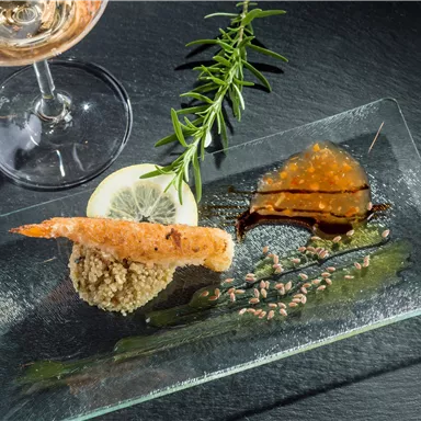 Ein kunstvoll angerichteter Teller mit einer Krabbe, Zitronenscheibe und Soßen. Im Hintergrund steht ein Glas Roséwein und ein Rosmarinzweig.