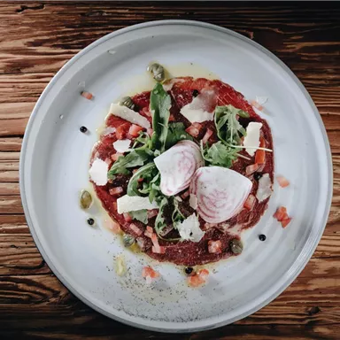 Ein appetitliches Gericht auf einem weißen Teller, bestehend aus rohem Fleisch, Salat und verschiedenen Toppings. Der Hintergrund ist aus hellem Holz.
