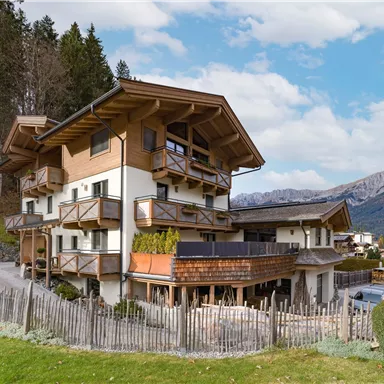 Ein charmantes Holzhaus in den Bergen mit mehreren Balkonen. Die Umgebung ist von grünen Wiesen und Bäumen umgeben.