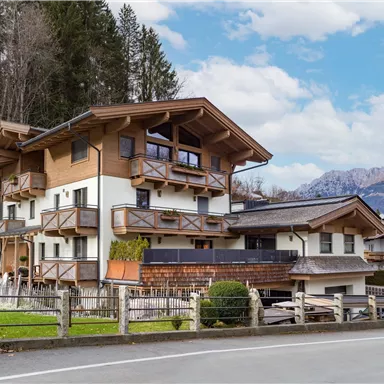 Ein modernes, alpines Haus mit mehreren Balkonen und einer Holzverkleidung. Im Hintergrund sind sanfte Berge und ein klarer Himmel zu sehen.