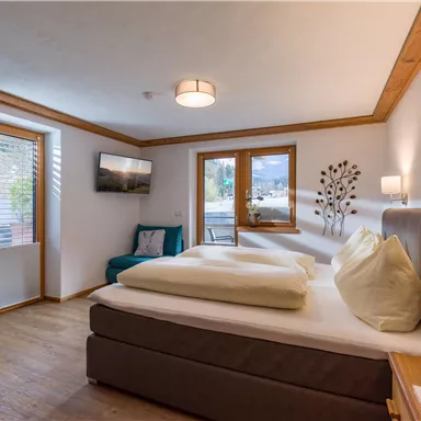 Ein gemütliches Schlafzimmer mit einem großen Bett und hellem Holzdekor. Fenster lassen viel Tageslicht herein, und eine bequeme Couch ergänzt den Raum.