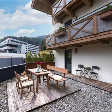 Ein gemütlicher Außenbereich mit Holzterrasse, Tisch und Stühlen. Im Hintergrund sind Berge und moderne Gebäude zu sehen.