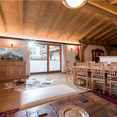 Ein helles Wohnzimmer mit Holzverkleidung und großen Fenstern. Es gibt einen Essbereich mit einem Holztisch und Stühlen sowie einen gemütlichen Teppich.