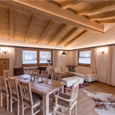 Ein gemütliches Wohnzimmer mit Holzdecke und großem Esstisch. Die Fenster lassen viel Licht herein und die Möbel sind modern und einladend.