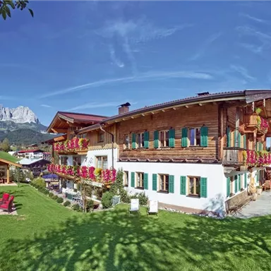 Ein charmantes Holzhaus mit bunten Blumenbalkonen, umgeben von grünem Rasen. Im Hintergrund sind majestätische Berge und ein klarer Himmel zu sehen.
