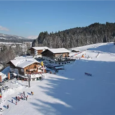 Eine schneebedeckte Landschaft mit gemütlichen Hütten und Menschen, die Ski fahren. Der klare blaue Himmel und die bewaldeten Berge schaffen eine idyllische Winteratmosphäre.