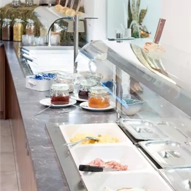 Ein Buffet mit verschiedenen Speisen, darunter Eier, Schinken und Marmelade. Der Esstisch ist hell und sauber gestaltet.