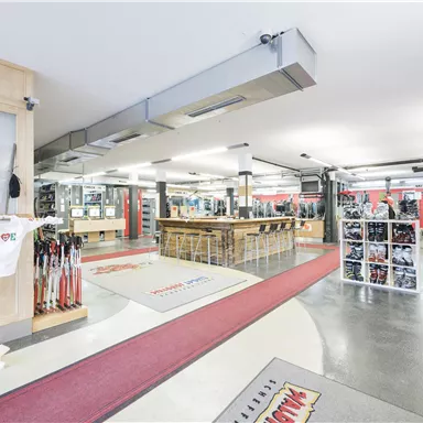 Ein moderner Verkaufsraum mit Sportartikeln und einer Bar. Die Wände sind mit Regalen und Spiegeln dekoriert, während der Boden mit rotem Teppich akzentuiert ist.