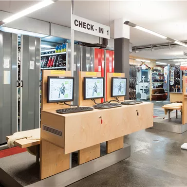 Ein modernes Check-in-Bereich mit mehreren Computerstationen. Im Hintergrund sind Regale mit Sportausrüstung sichtbar.