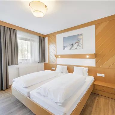 Ein helles Schlafzimmer mit einem großen Doppelbett und Holzdekoren. Große Fenster sorgen für viel Tageslicht und eine gemütliche Atmosphäre.