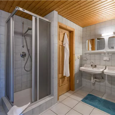 A modern bathroom with a shower and two sinks. The walls are tiled and there is a wooden ceiling covering.