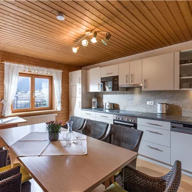 A modern kitchen with a wooden ceiling and white cabinets. A dining table with chairs is in the foreground, accompanied by a decorative plant.