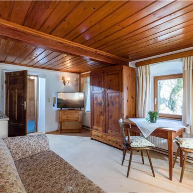 A cozy living room with a wooden ceiling and a seating area. There is a table, chairs, and a television, as well as plenty of daylight through the window.