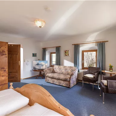 A cozy room with a double bed and a comfortable sofa. There are windows with curtains and a small table with chairs.