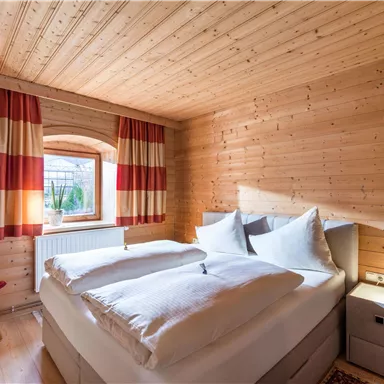 A cozy bedroom with wood paneling and large windows. The bed is dressed with white pillows and blankets, and the curtains have a red and white striped pattern.