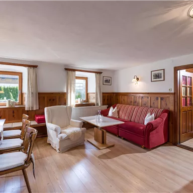A cozy living space with wooden paneling and large windows. A red sofa and a dining table with chairs invite relaxation.