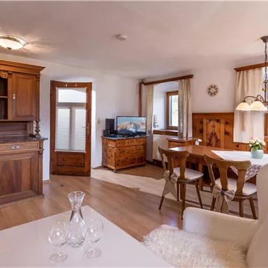 A cozy apartment with wooden furniture and a dining table. Large windows let in a lot of light into the room.