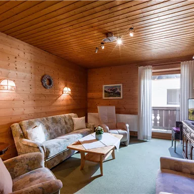 A cozy living room with wooden walls and warm lighting. There are comfortable sofas and a small table, perfect for relaxing.