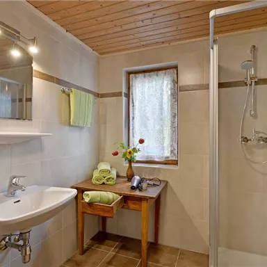 Ein modernes Badezimmer mit einer Dusche, einem Waschbecken und einem Holzregal. Es gibt frische Handtücher und eine kleine Blumendekoration auf dem Tisch.