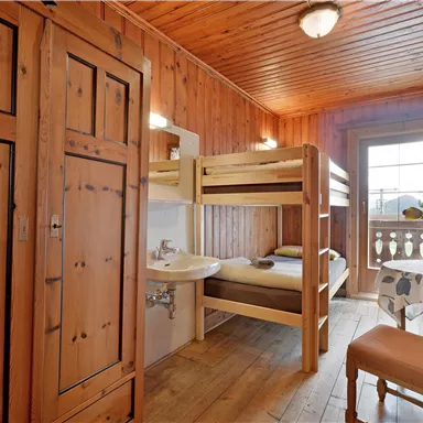 Ein gemütliches Zimmer aus Holz mit einem Etagenbett und einem Schreibtisch. Das Fenster bietet einen Blick auf den Balkon und die umliegende Landschaft.