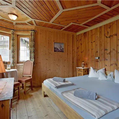 Ein gemütliches Zimmer mit Holzverkleidung und einem großen Bett. Ein Tisch mit Stuhl und ein sanftes Licht schaffen eine einladende Atmosphäre.