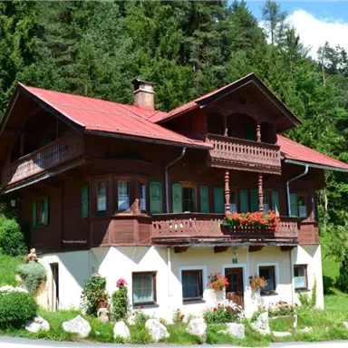 Ein traditionelles Holzhaus mit einem roten Dach und blühenden Blumen. Es steht inmitten von grünen Bäumen und einer malerischen Landschaft.