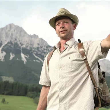 Ein Mann steht entspannt an einem Baum und schaut in die Ferne. Im Hintergrund sind beeindruckende Berge und eine grüne Landschaft zu sehen.