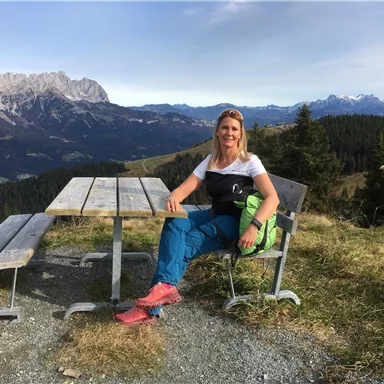 A woman is sitting at a table looking at the mountains. The landscape is characterized by trees and the Alps.