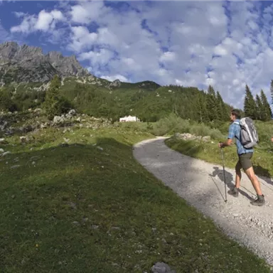 Two hikers are walking on a narrow path through a green landscape. In the background, there are high mountains and a blue sky.