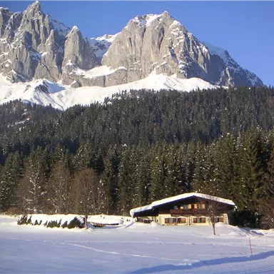 Ein schneebedecktes Tal mit einer charmanten Berghütte im Vordergrund. Im Hintergrund erheben sich majestätische Berge unter einem klaren blauen Himmel.
