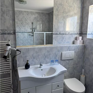 Ein modernes Badezimmer mit einer weißen Waschbeckenzeile und einem großen Spiegel. Die Wände sind hell gefliest und es gibt eine Dusche sowie Hygieneartikel.